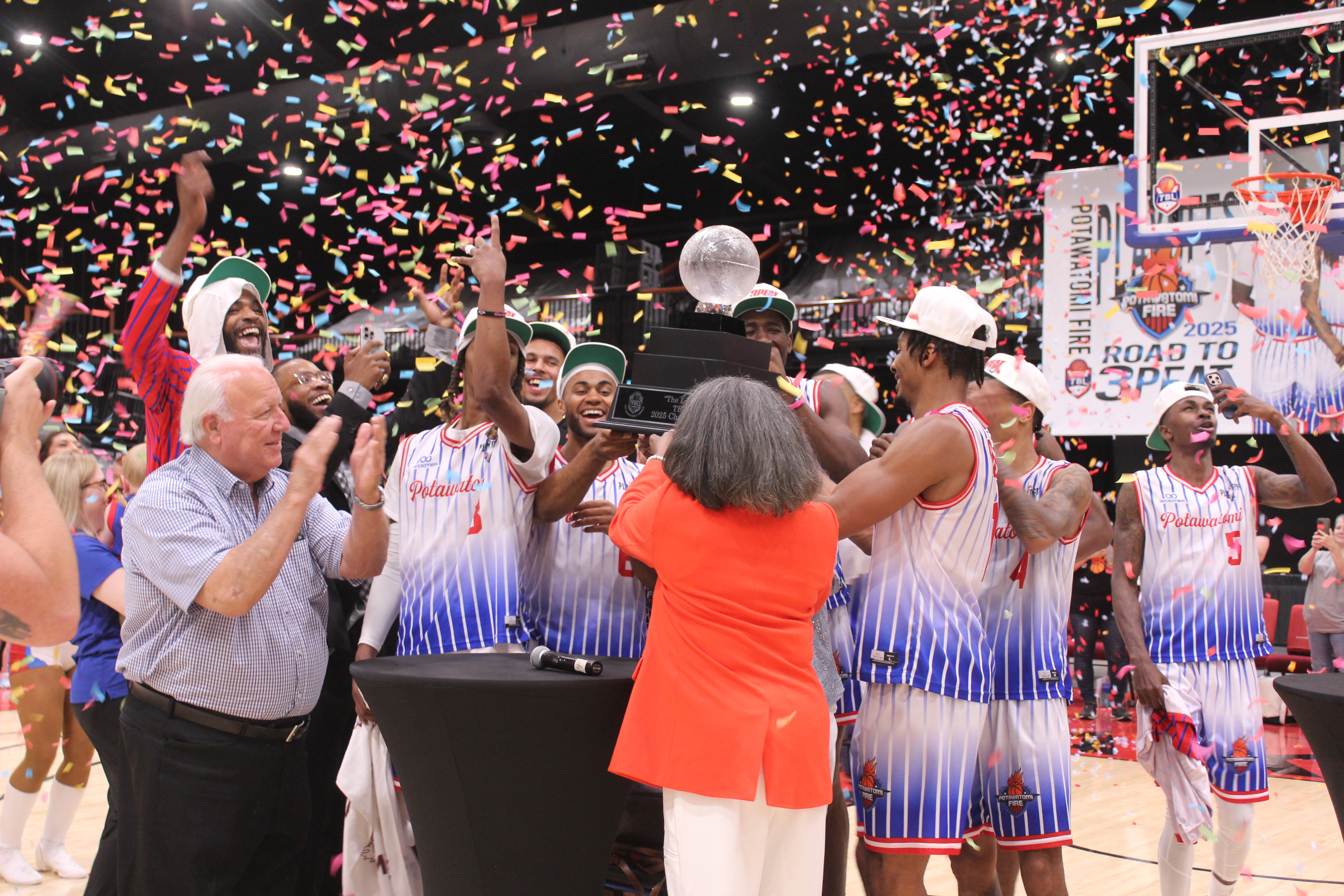 The Potawatomi Fire Team, along with Citizen Potawatomi Nation Chairman John 'Rocky' Barrett, The Basketball League CEO Evelyn Magley (back to camera) and Lillie Mae Greer (hidden by her daughter), celebrate their Three-Peat by lifting 'The Lillie' TBL 2025 Champion trophy high, amid confetti on June 23 at FireLake Arena. This is the third year in a row they have won this national title, the first in the TBL to do so. Their record for the season is 27-1, and 8-0 for the playoffs.