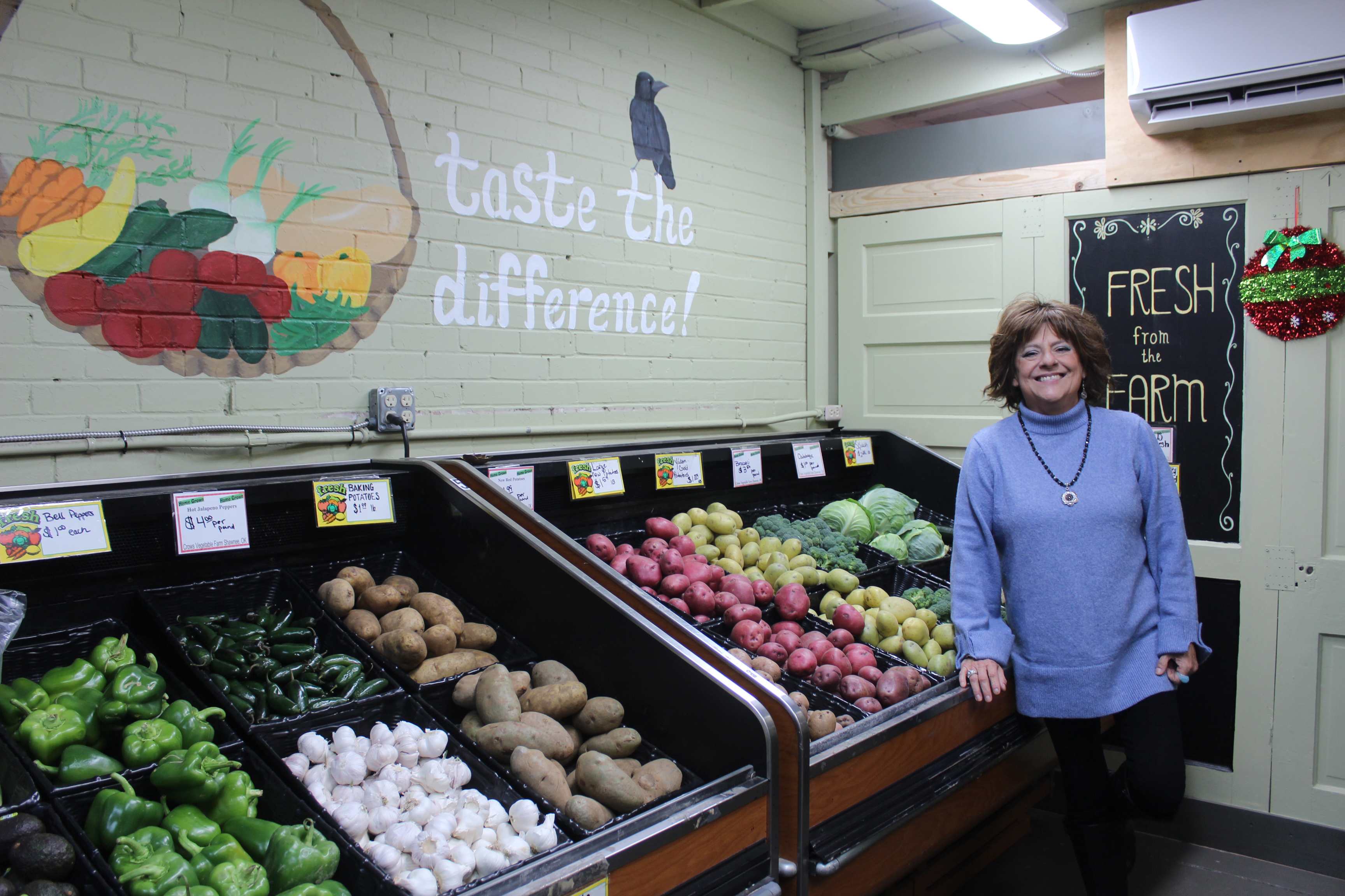 Crows Farm Markets Co-Owner Claudia Crow poses next to the vegetables grown on their farm by her husband, Ricky Crow. C. Crow said a woman who worked at the market painted the mural, and they stand by the statement that home-grown vegetables have more flavor than those from big-box grocery stores. Crows Farm Market in Shawnee will close at the end of this month after 25 years under the Crows' ownership. Natasha Dunagan, Countywide & Sun