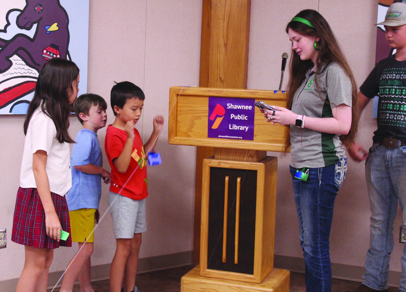 State 4-H STEM Innovator Tori Nelms watches the stopwatch on her phone as Nina Tomlin, 9; Barrett Gates, 8; and Norimi Tomlin, 7; all of Shawnee, test their zip line container, which holds a ping pong ball, during the 'Build It: Zip Line Zoom' program at the Shawnee Public Library on July 21. Children worked together to build their containers, then tested them on six already-made zip lines made of cotton string or fishing line. Fellow Innovators Madison Maddox & Tony Nelms, plus 4-H Educator Kacie Jasper.