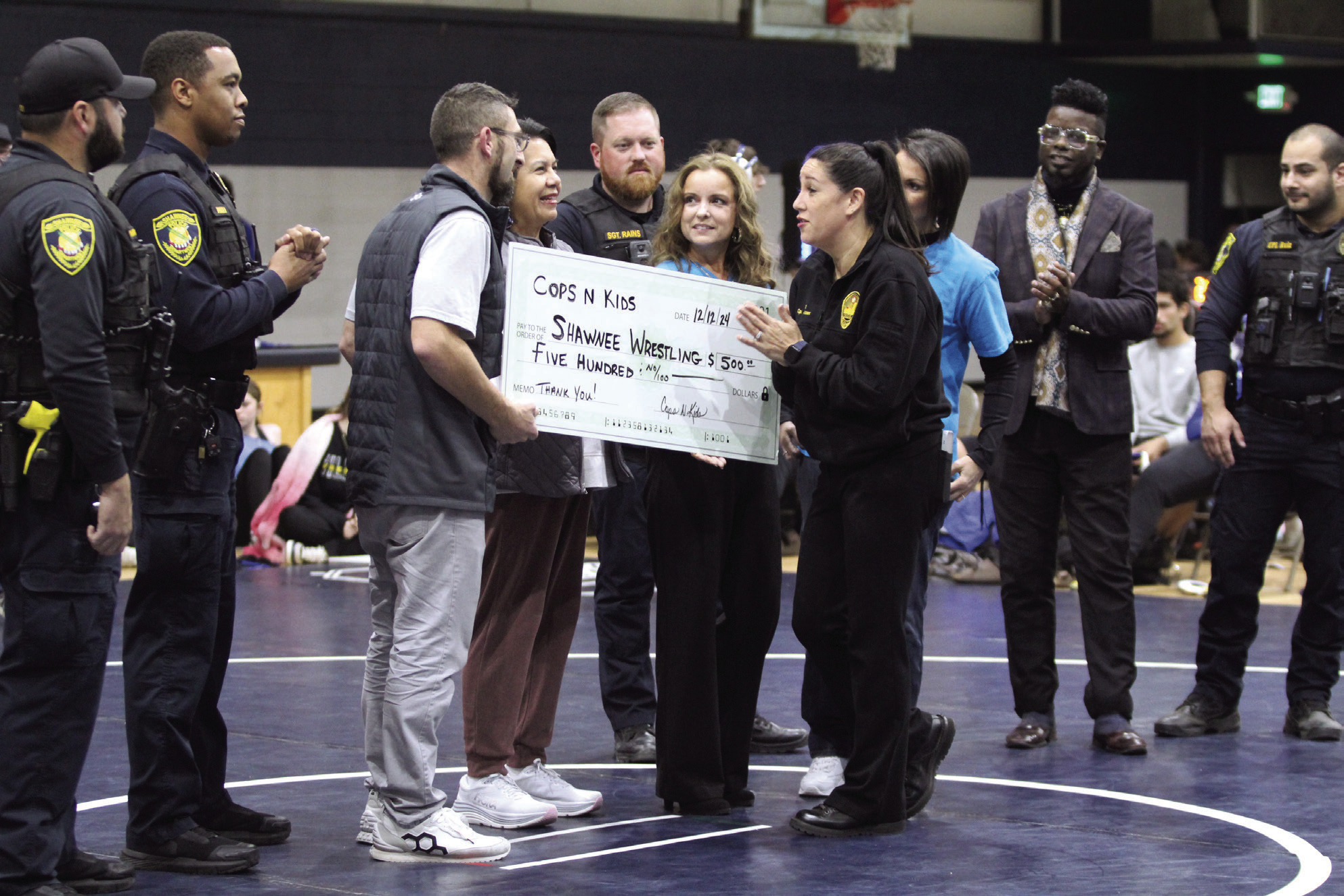 Vivian Lozano, the founder of Cops n' Kids, presents a check from the Cops n' Kids Board for $500 to Shawnee Wrestling Coach Andrew McCune for the Shawnee Wrestling Program during the home dual on Dec. 12. Lozano said the board plans to donate to different causes in the community every year. Present for the presentation, not in order, were board members Tara Andrews, Sally Coleman, Lisa Watson and Jeff Chamblain, as well as Jamelle Payne, Sgt. Rains, Cpl. Ruiz, Cpl. Price and PTL Arispe.