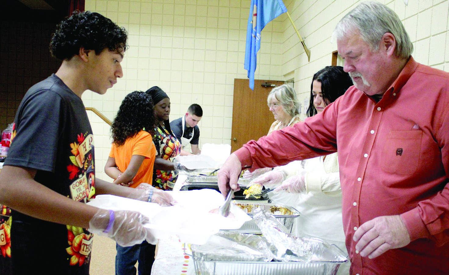 Randy Gilbert adds turkey to the dinners held by volunteer Vergil Brawner, 14, at the 19th Annual Tecumseh Family Thanksgiving Dinner, held at Tecumseh City Hall Auditorium on Nov. 28. Brawner's sister, Lyric Brawner, 10, and mother, Maud Police Chief Doneisha Brawner, shown in background, also volunteered. Also shown are Mason Blumenthal, Melissa Hewett and Annie Jo Gilbert. See full cutline below.
