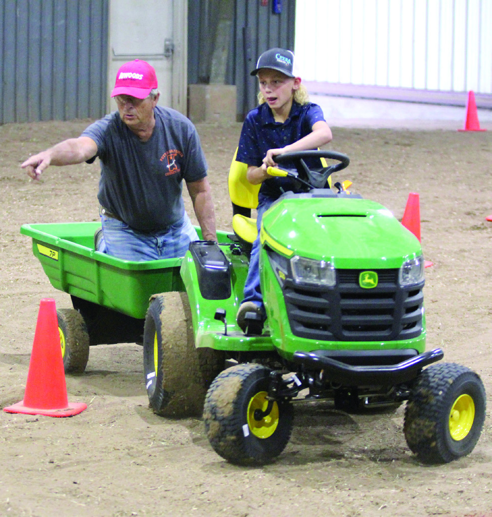 Dace Dockrey, age 9, drives a garden tractor through the obstacle course of the Peewee and Midget Tractor Operator Contest, as Fair Board Member Lynn Marlow, points to where he needs to go, on Sept. 6 at the Pottawatomie County Free Fair. File photo