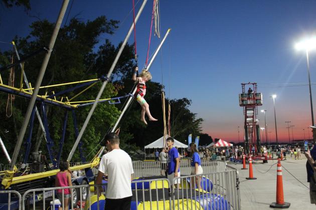 As the sun sets on Aug. 13, a young girl jumps high above a trampoline with the help of bungee cords and a harness. To the right, the zip line can be seen. This was the first year for the FireLake Fireflight Balloon Festival to offer carnival wristbands for Extreme Inflatables rides. Countywide &amp; Sun/Natasha Dunagan