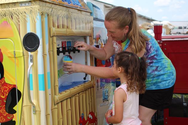 Amy Sanders, co-owner of the Kona Ice franchise in Shawnee, helps Violetta Whitley, 3, of Pink, to add flavoring to her snowcone during the Farming Heritage Festival at Shawnee Feed on June 17. Co-owner Aron Sanders said they also go to local schools each week and give a portion of the profits to the school. Nationwide, he said, Kona Ice gave $135 million back to communities last year. Countywide &amp; Sun/Natasha Dunagan