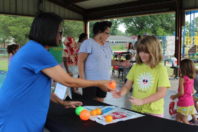 Lisa Watson hands a ball to Lily McCracken, 9, of Tecumseh, at the Gateway to Prevention and Recovery (TSET) Healthy Living Program booth on July 25. Watson and Lisa Smith, center, also offered mandarin oranges, bottled water and cooling towels at the Tecumseh Pioneer Library "Party at the Park," held at Slick Humphrey Park. Countywide &amp; Sun/Natasha Dunagan