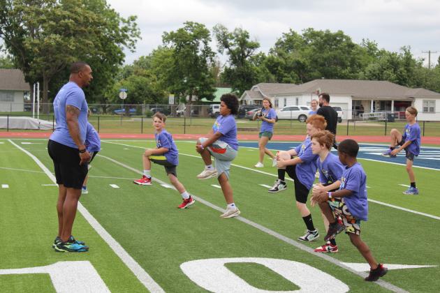 Former NFL safety Shamiel Gary directs elementary students in a warm-up with knee hugs during his first of two football clinics on May 29 at Jim Thorpe Stadium in Shawnee. There were more than 40 children in this clinic, sponsored by Pottawatomie Go (POGO) for Health Week, and there were over 20 middle school students in the second football clinic that day. Several Shawnee High School football players and their coaches helped with both clinics.