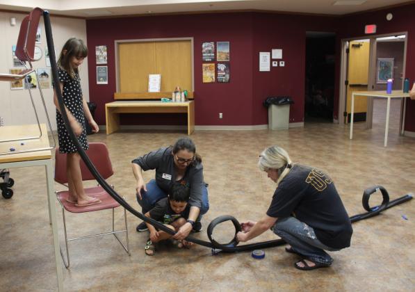 Rhiannon Hayes, 8, on chair; Colton Hayes, 5; both of Shawnee; 4-H Educator Kacie Jasper; and the childrens' mother Margie Stanton work on a 3-loop roller coaster during the "Build It: Roller Coaster" event on June 14. They used insulation tubing, masking tape and a cup to catch the marble. Jasper was assisted by State 4-H STEM Innovators Grace Hobbs and Madison Maddox. The next Build It event will be "Ready, Set, Blast Off!" on June 30 at 2 pm.