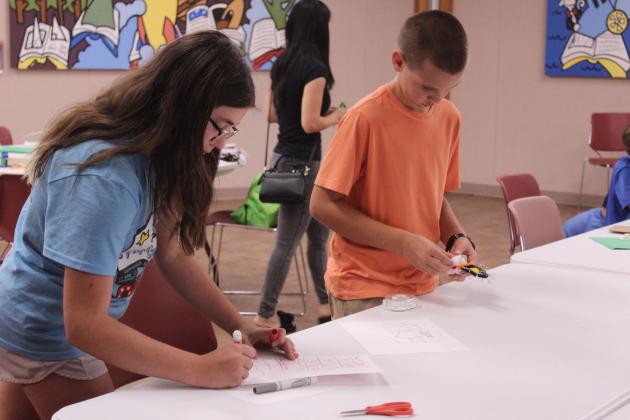 Pierce Muir, right, 10, of Tecumseh, adds tape to their zip line container, as Ava Zarrella, 12, of Shawnee, keeps track of how many seconds it ran on each zip line using a chart, during the "Build It: Zip Line Zoom" program at the Shawnee Public Library on July 21. Children worked together to build their containers, then tested them on six already-made zip lines made of cotton string or fishing line.