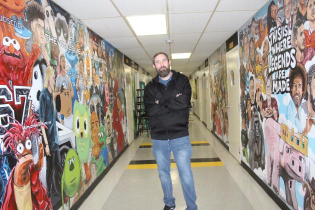 Tecumseh Middle School Art Teacher Kelly Renken stands in the hallway outside his room between the Massive Meme Memorial and the Wall of Legends. In the past year, he has painted 85 memes and 56 legends in the TMS North Building, and he posts his work on YouTube and TikTok, with 52.3K subscribers so far.
