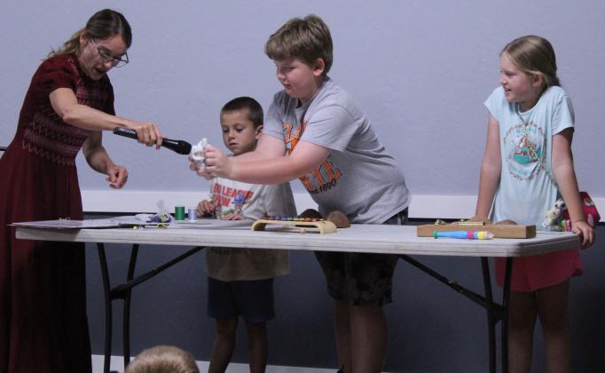 Luke Elliott, 9, of McLoud, crumples paper to make the sound effect for fire, as two other volunteers look on, and Erin Yeaman of Norman reads 'The Three Little Pigs.' Yeaman was showing how ordinary items can 'make music.' She also played her cello along to three children's books and let everyone try their hands at playing the cello during the 'Tunes and Tales with Erin Yeaman' McLoud Public Library program, held on June 18 at the Ray Large American Legion Post 244. See full caption below.