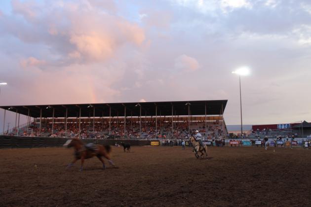 A rainbow ends at the Grandstands of the IFYR, as volunteers and contestants go about the business of steer wrestling in Arena #1 during the championship round on the evening of July 17.