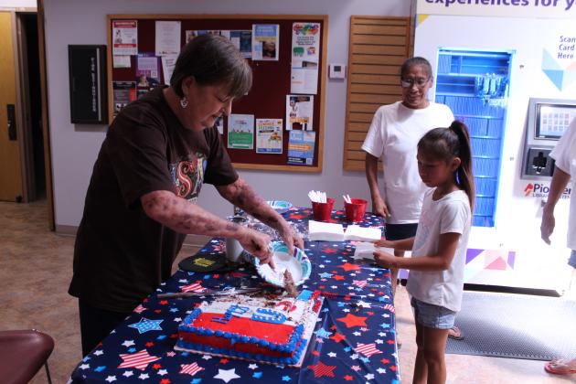 Shawnee Public Library Branch Manager Mary Sims serves "Happy Birthday, USA!" birthday cake to Jasmin Ataddlety, 7, of Shawnee, as Clarissa Ataddlety looks on, during the third day of 250th USA anniversary celebrations in the library lobby on July 1. They offered popcorn and ice cream sandwiches on June 29 and the Snow Shak offered free snow cones at the library on June 30. They also had a balloon arch and their finished community tile artwork on display, available to the public for a photo backdrop.