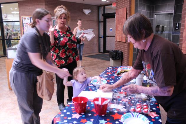 Shawnee Public Library Branch Manager Mary Sims offers napkins, along with their USA birthday cake, to Alyssa Wissinger and Brielle Jackson, 2, both of Shawnee, as Deedee Thomas of Tecumseh looks on, during the third day of 250th USA anniversary celebrations in the library lobby on July 1. They offered popcorn and ice cream sandwiches on June 29, and the Snow Shak offered free snow cones at the library on June 30.