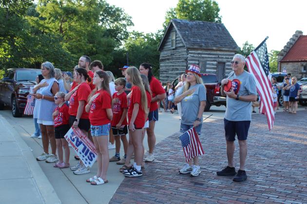Attendees, including Tanner Silas supporters, former Shawnee Mayor Ed Bolt and Phyllis Bolt, salute during the playing of the "Star Spangled Banner" before the Shawnee Independence Day Parade on July 4. Participants in the parade gathered at the Pottawatomie County Museum (Paul and Ann Milburn Center) at 7 am. The Oklahoma Jazz Preserve performed and led the parade. The historic Beard Cabin is shown in the background.
