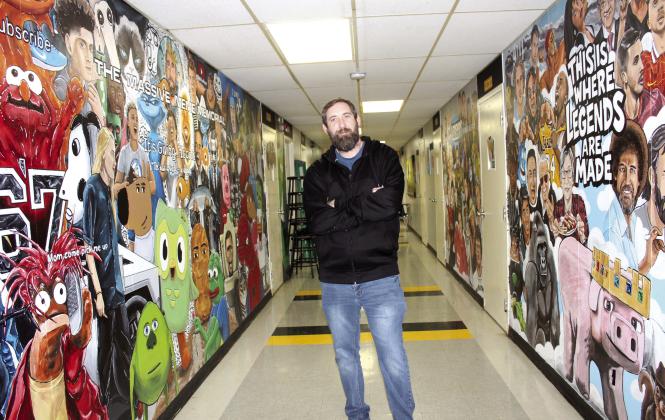 Tecumseh Middle School Art Teacher Kelly Renken stands in the hallway outside his room between the Massive Meme Memorial and the Wall of Legends. In the past year, he has painted 85 memes and 56 legends in the TMS North Building, and he posts his work on YouTube and TikTok, with 52.3K subscribers so far.