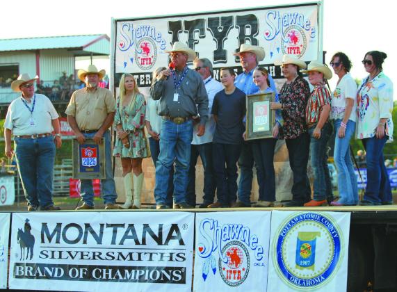 On July 9, IPRA/IFYR General Manager Dale Yerigan speaks about Randy and Suzanne Gilbert after presenting them with a framed 2025 IFYR logo with engraved brass plate in honor of their 33 years of volunteer service to the rodeo. To the right, the Gilberts are shown with their family: Legend Baker, Taycie Baker, Bryar Baker, Dustie Baker and Annie Jo Gilbert. Also shown, from left, are State Representative Dell Kerbs, Greg Rush, Melissa Dennis, Abby Thompson (hidden) and Shawnee Mayor Eric Stephens.