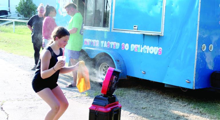 In 100-degree temperatures, Laramie Dombrowski, 12, of Shawnee, cools down with a misting fan and a snow cone, as The Snow Shak - owned by her mother, April Dombrowski, shown in background far left - gives away snow cones.