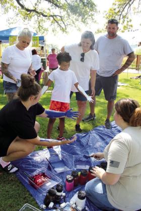 Kayla Beal, kneeling left, and Exzavier Beal, 7, both of Shawnee, look at their arms and hands after Olivia Rawlins, of Tecumseh, kneeling right, finished painting them so they could create a “Forever Hug” during the First Annual Hug-A-Thon at Woodland Veterans Park on Aug. 3. Also shown, standing from left, are Rita Beal, Alexis McCool and Arthur McCool. Countywide &amp; Sun/Natasha Dunagan
