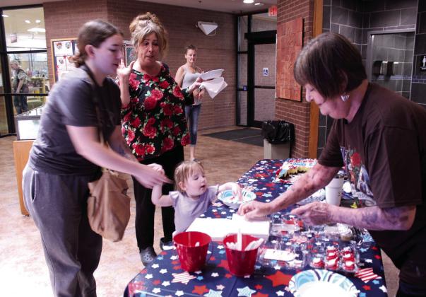 Shawnee Public Library Branch Manager Mary Sims offers napkins, along with their USA birthday cake, to Alyssa Wissinger and Brielle Jackson, 2, both of Shawnee, as Deedee Thomas of Tecumseh looks on, during the third day of 250th USA anniversary celebrations in the library lobby on July 1. They offered popcorn and ice cream sandwiches on June 29, and the Snow Shak offered free snow cones at the library on June 30.