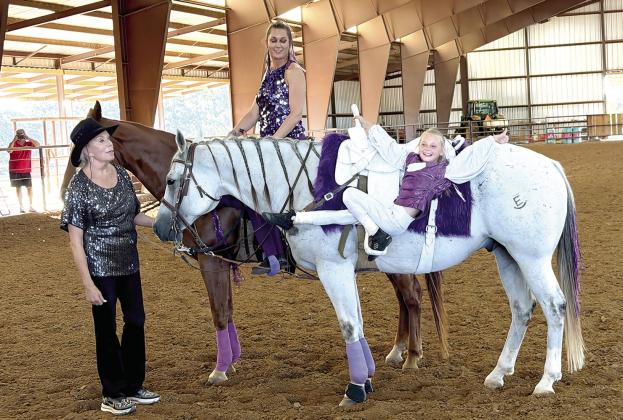 Jensen Dockery, eight-year-old daughter of Shane and Mandy Dockery, demonstrates one of several tricks she performed Saturday evening at the Shootin' &amp; Scootin' for Justice event. Jensen began trick-riding lessons just four weeks prior to the event. Also pictured with her are Penny Treeby-Hudgens and Desireé Treeby on horseback. Suzie Campbell, Countywide &amp; Sun