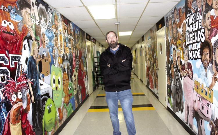 Tecumseh Middle School Art Teacher Kelly Renken stands in the hallway outside his room between the Massive Meme Memorial and the Wall of Legends. In the past year, he has painted 85 memes and 56 legends in the TMS North Building, and he posts his work on YouTube and TikTok, with 52.3K subscribers so far.