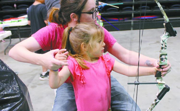 Janilee Marcus, with the Outdoor Nation Expo Youth Archery booth, helps Savannah Morris, 4, of Washington, to shoot a bow for the first time at FireLake Arena on Aug. 9. This is the first year, since the ONE began in 2017, for it to be presented by the Citizen Potawatomi Nation. They also had the Fetch n' Fish Dog Show and Darkwater Adventures Target Bowfishing, as well as an assortment of vendors. Natasha Dunagan, Countywide & Sun